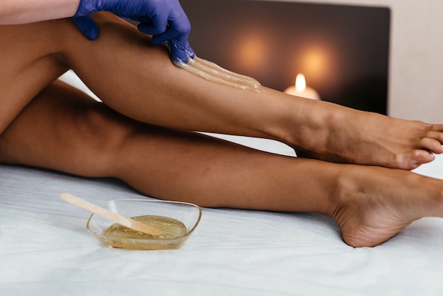 Manos de primer plano de cosmetóloga en guantes azules aplicando pasta para depilación con azúcar en la pierna femenina, procedimiento de belleza de depilación.