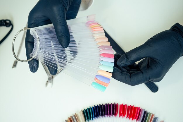 Manos de primer plano de maestro profesional manicurista de mujer de manicura en una chaqueta blanca y guantes de goma negros es elegir una paleta de colores de esmalte de uñas en un salón de belleza spa.