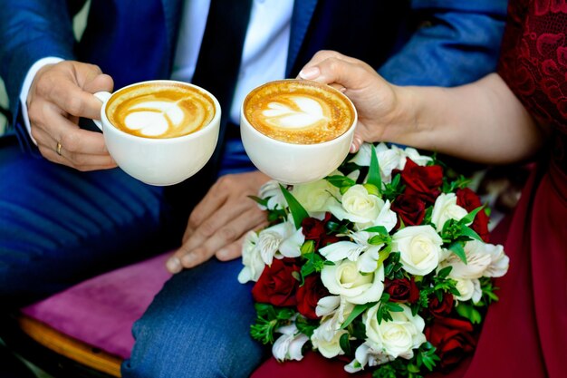 Las manos de los recién casados están sosteniendo tazas de café un ramo en el fondo