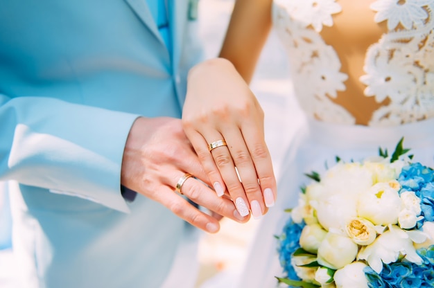 Manos de recién casados con hermosos anillos de oro, primer plano. Vestido de dama de honor blanco, ramo, manicura elegante. Ceremonia de boda perfecta.