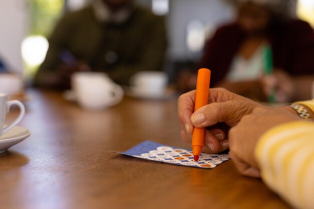 Manos recortadas de una anciana birracial marcando números en una tarjeta de bingo sobre una mesa en un hogar de ancianos. Rotulador, suerte, juego de ocio, inalterado, apoyo, vida asistida, concepto de jubilación.