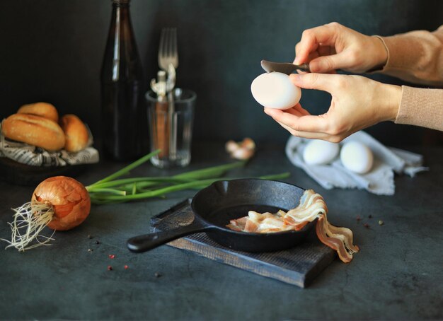 Las manos rompen un huevo sobre una sartén en la mesa de la cocina en casa Cocinar huevos revueltos y tocino