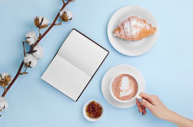 Manos sosteniendo café y cuaderno con croissant y rama de algodón