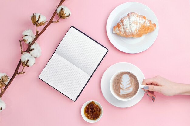 Manos sosteniendo café y cuaderno con croissant y rama de algodón