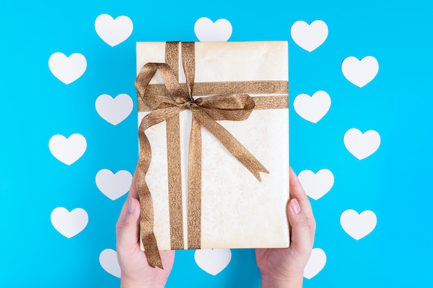 manos sosteniendo una caja de regalo amarilla atada con una cinta marrón brillante sobre corazones blancos colocados en azul