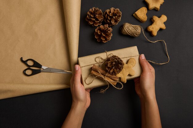 Foto manos sosteniendo una caja de regalo de navidad hecha a mano envuelta en papel artesanal marrón decorado con piñas y canela sobre una superficie negra con un hombre de pan de jengibre y galletas, cuerda y tijeras. endecha plana