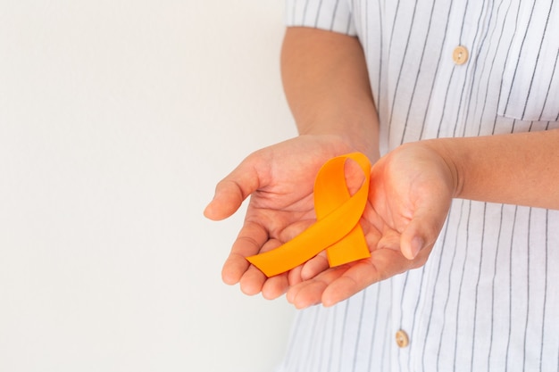 Manos sosteniendo cinta de color naranja sobre tela blanca con espacio de copia. Concienciación sobre el cáncer de riñón, Enfermedad de leucemia, Concienciación sobre el cáncer de piel, Día mundial contra el cáncer. Concepto de salud u hospital y seguro.