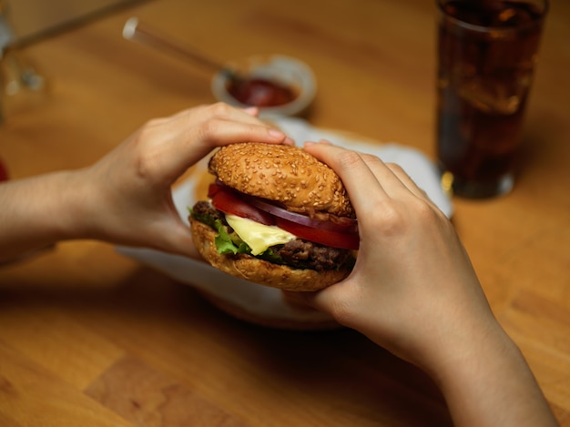 Manos sosteniendo una deliciosa hamburguesa de tocino fresca lista para comer en la mesa con salsa de tomate y bebida