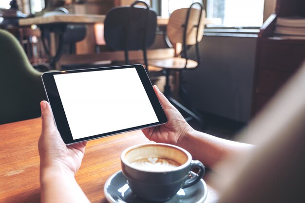 Manos sosteniendo negro tablet pc con pantalla blanca en blanco con taza de café