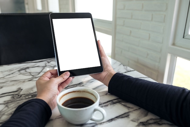 manos sosteniendo la pc de la tableta negro con pantalla blanca en blanco y taza de café en la mesa de café