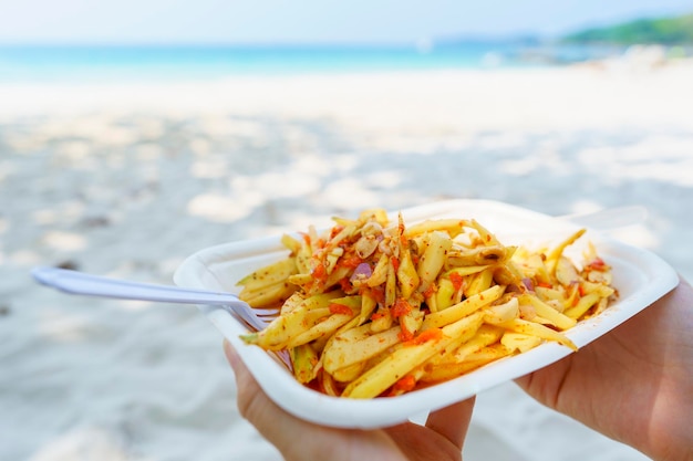 Foto manos sosteniendo un plato de deliciosa ensalada de papaya con fondo de mar y playa comida de playa de tailandia