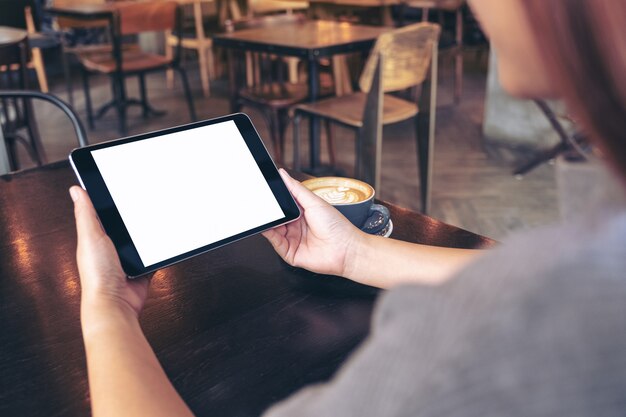Manos sosteniendo tablet pc negro con una pantalla en blanco con taza de café