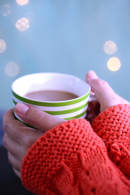 Manos sosteniendo la taza de bebida caliente, primer plano, sobre fondo brillante