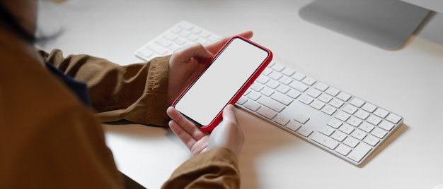 Manos sosteniendo el teléfono inteligente de pantalla en blanco sobre el teclado de la computadora en el escritorio de oficina