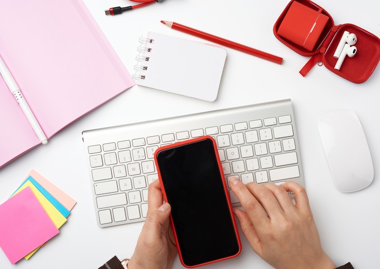 Foto manos sosteniendo un teléfono inteligente con una pantalla negra en blanco sobre un escritorio blanco con un teclado, mouse de computadora, cuaderno y pegatinas de colores