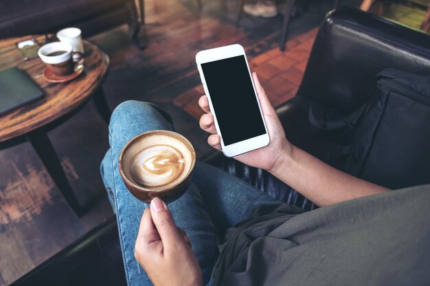 Manos sosteniendo un teléfono móvil blanco con pantalla en blanco en el muslo y la taza de café