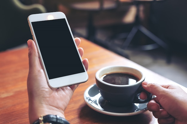 Manos sosteniendo un teléfono móvil blanco con pantalla en blanco negro mientras bebe café en la cafetería moderna