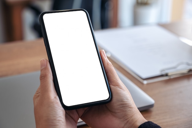 Manos sosteniendo un teléfono móvil negro con pantalla en blanco en blanco con un portátil en la mesa de madera en la oficina