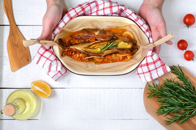 Las manos sostienen el pescado recién cocido. Verduras, hierbas aromáticas, aceite de oliva en una mesa de madera blanca. Vista superior