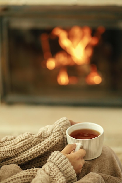 Foto manos en un suéter acogedor sosteniendo una taza de té caliente en la chimenea ardiente cerca de la calefacción hygge de otoño
