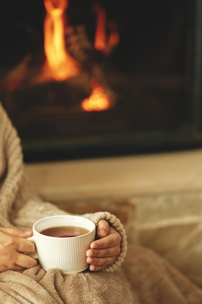 Manos en suéter acogedor sosteniendo una taza de té caliente en el fondo de la chimenea en llamas cerrar hygge