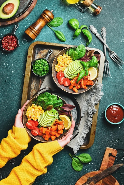 Las manos en un suéter amarillo sostienen un cuenco de ensalada calabaza aguacate maíz y tomates concepto de alimentación limpia vista superior