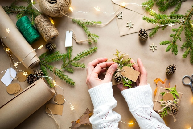 Manos en un suéter blanco empaquetan un regalo para Navidad y año nuevo en materiales ecológicos: papel kraft, ramas de abeto vivo, conos, cordel. Etiquetas con mock up, decoración natural, hechas a mano, bricolaje. Actitud festiva