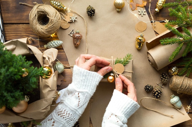 Manos en un suéter blanco empaquetan un regalo para Navidad y año nuevo en materiales ecológicos: papel kraft, ramas de abeto vivo, conos, cordel. Etiquetas con mock up, decoración natural, hechas a mano, bricolaje. Actitud festiva