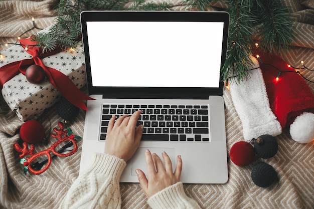 Manos trabajando en una computadora portátil con pantalla en blanco en una cama acogedora con elegantes adornos navideños presentes