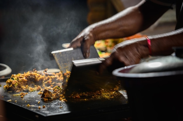 Manos del vendedor de kottu roti haciendo kottu encima de una placa de parrilla de acero caliente