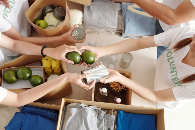 Foto manos de voluntarios que pasan comida enlatada y frutas cítricas verdes cuando empacan cajas para refugiados