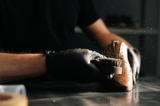 Las manos de un zapatero con guantes negros quitan el polvo del cepillo de zapatos en una zapatería artesanal oscura. Concepto de trabajo de reparación y restauración artesanal de zapatero en taller de reparación de calzado.
