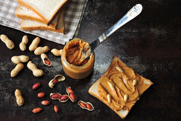 Manteiga de amendoim em uma jarra, faca, amendoim e torrada de trigo fresco com manteiga de amendoim. Lanche vegetariano ou vegan. Conceito de pequeno-almoço saudável.