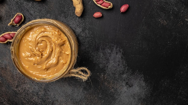 Manteiga de amendoim ou pasta em uma jarra aberta e amendoim na casca espalhados em um lugar de receita de menu de banner de fundo escuro para exibição superior de texto