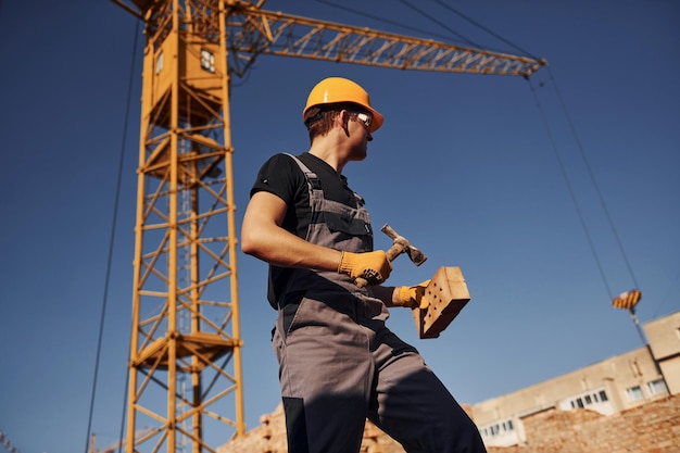 Mantém tijolo e ferramenta nas mãos Trabalhador da construção em uniforme e equipamento de segurança tem trabalho na construção