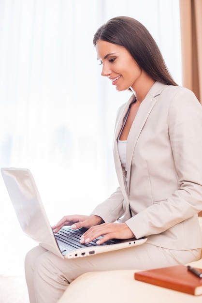 Mantenerse conectado. Vista lateral de la hermosa joven empresaria en traje trabajando en la computadora portátil y sonriendo mientras está sentado en la cama