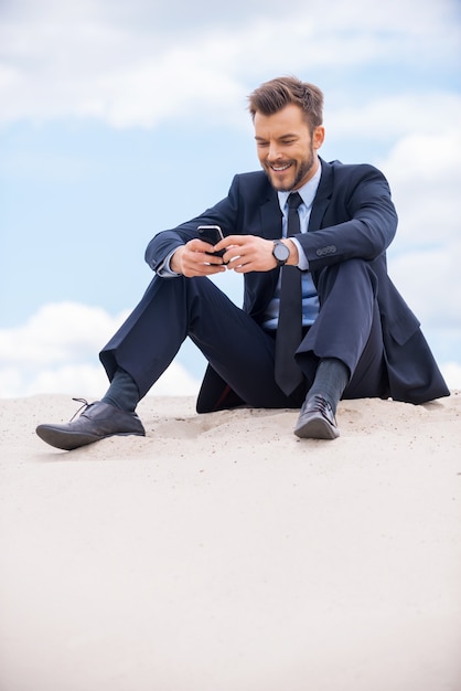 Mantenerse en contacto. Alegre joven empresario mirando su teléfono móvil y sonriendo mientras está sentado en la arena y contra el cielo azul