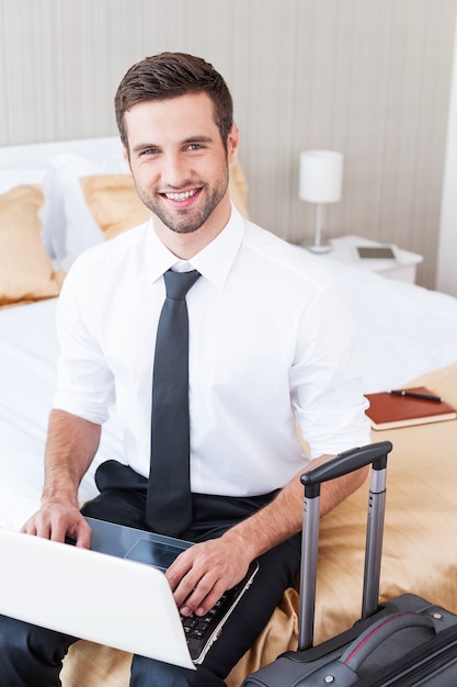 Mantenerse en contacto en cualquier momento y en cualquier lugar. Vista superior del apuesto joven en camisa y corbata trabajando en una computadora portátil y sonriendo mientras está sentado en la cama en la habitación del hotel