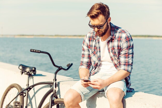 Mantenerse en contacto mientras está de vacaciones. Hombre joven pensativo que sostiene el teléfono móvil mientras está sentado en el parapeto cerca de su bicicleta