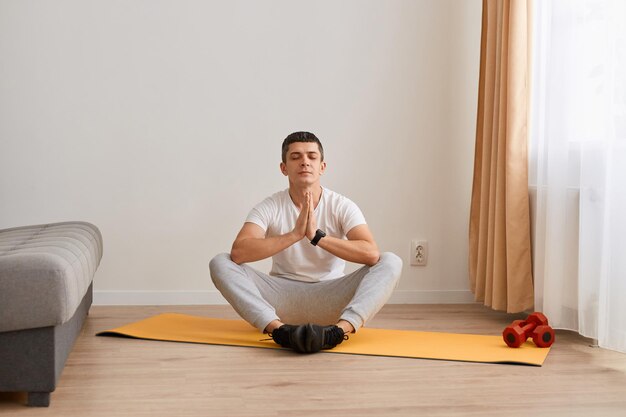 Mantenga la calma Hombre caucásico deportivo feliz meditando con un entrenador en línea a través de una conexión de computadora portátil usando una PC en la sala de estar chico sano sentado en la alfombra de yoga del piso en posición de loto