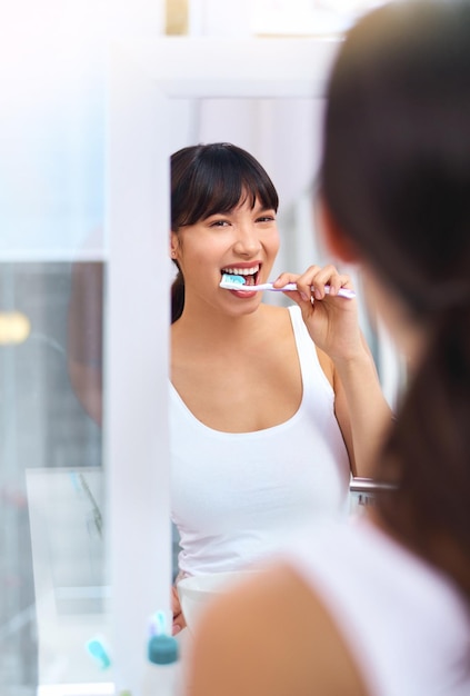 Manteniendo esas perlas blancas Foto de una joven alegre y atractiva cepillándose los dientes mientras mira su reflejo en un espejo en casa durante el día