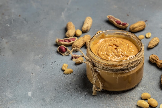 Mantequilla o pasta de cacahuete en un frasco abierto y cacahuetes en la cáscara esparcidos en un lugar de receta de menú de banner de fondo gris para la vista superior de texto