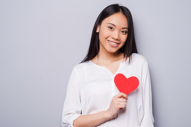 Manter o amor em seu coração. Bela jovem asiática segurando um cartão de dia dos namorados em forma de coração e sorrindo em pé contra um fundo cinza