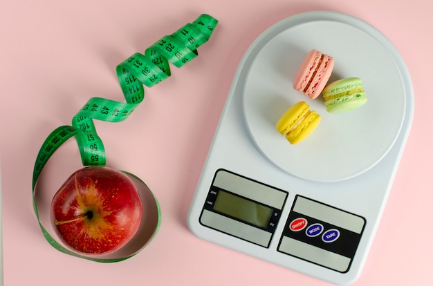 Manzana roja con cinta métrica verde, báscula de cocina digital con macarons en rosa