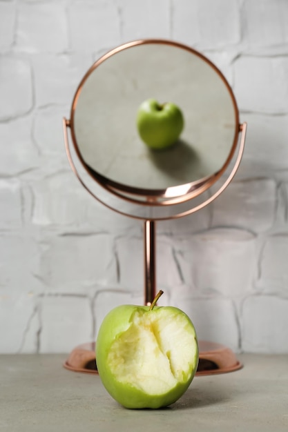 Manzana verde mordida cerca del espejo con reflejo de fruta entera en la mesa