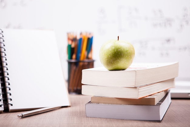 Manzana verde sobre una pila de libros junto a un cuaderno y lápices sobre la mesa con una pizarra blanca borrosa en la parte posterior. Concepto de escuela