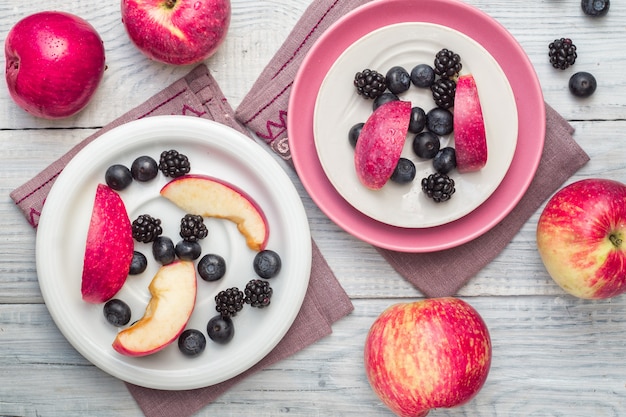 Manzanas rojas, moras y arándanos en placas sobre una mesa de madera blanca