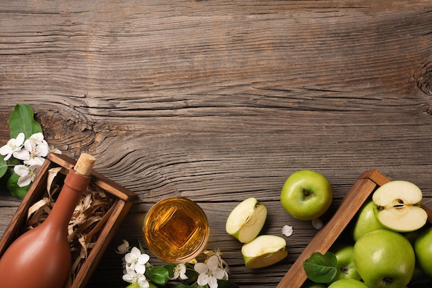 Manzanas verdes maduras en caja de madera con rama de flores blancas, vidrio y botella de sidra en una mesa de madera. Vista superior con espacio para su texto.