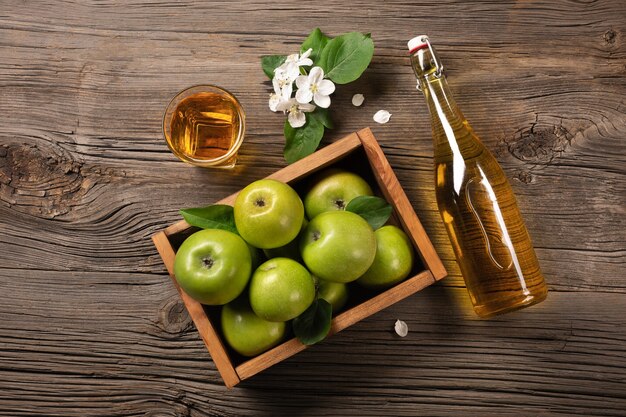 Manzanas verdes maduras en caja de madera con rama de flores blancas, vidrio y botella de sidra en una mesa de madera. Vista superior.