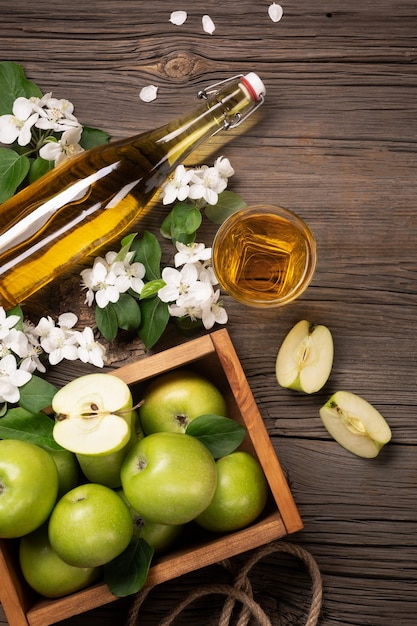 Manzanas verdes maduras en caja de madera con rama de flores blancas, vidrio y botella de sidra en una mesa de madera. Vista superior.